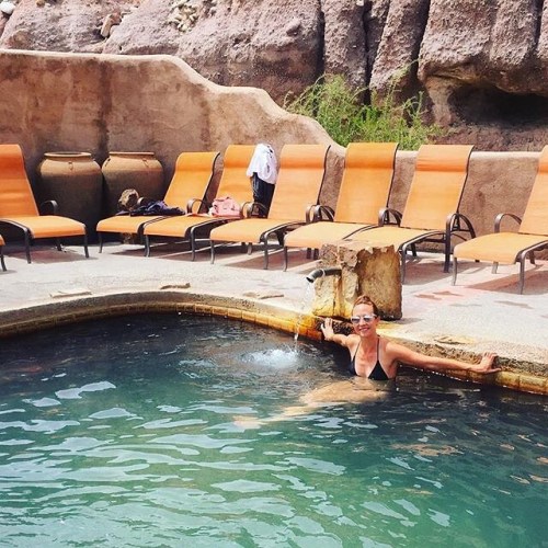 A person relaxes in a pool surrounded by lounge chairs and rocky terrain, enjoying a sunny day.