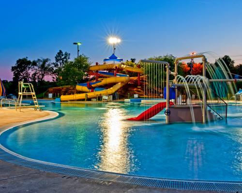 A vibrant water park at dusk featuring slides, a splash pad, and a swimming pool under bright lights.