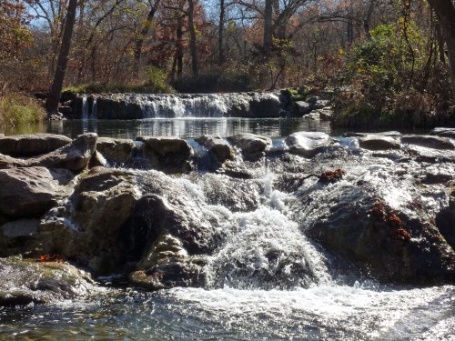 A serene waterfall cascades over rocks, surrounded by trees and autumn foliage under a clear blue sky.