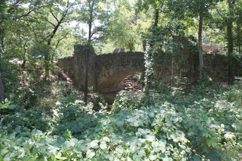 A stone bridge arches over a small stream, surrounded by lush greenery and trees in a wooded area.