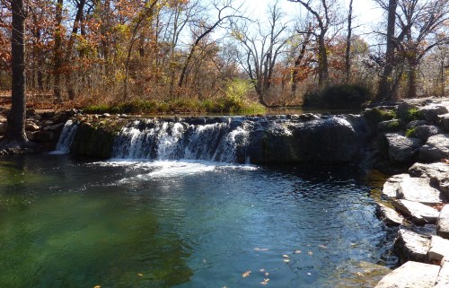 A serene waterfall cascades into a clear pool, surrounded by autumn trees and rocky banks.