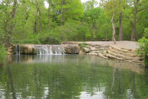 A serene landscape featuring a small waterfall cascading into a calm pond, surrounded by lush green trees.