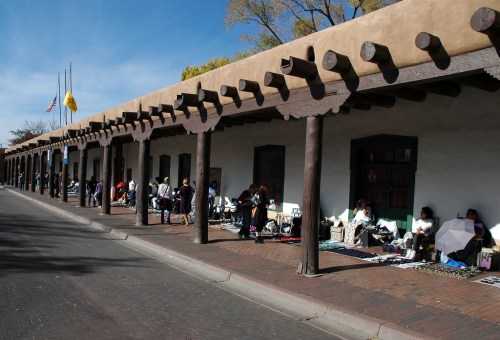 A row of adobe buildings with wooden beams, featuring vendors and shoppers along a sunny street.
