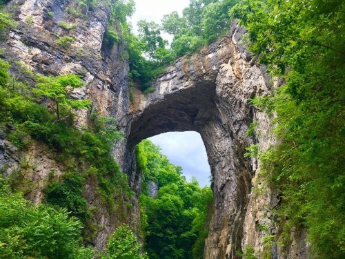 A natural stone arch surrounded by lush greenery and rocky cliffs under a cloudy sky.