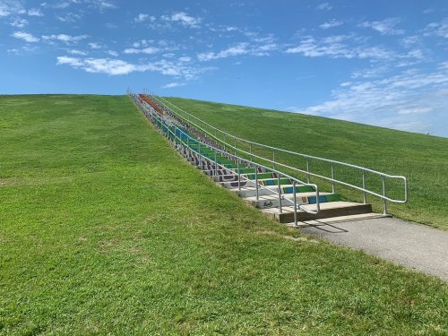 A colorful staircase leads up a grassy hill under a blue sky with scattered clouds.