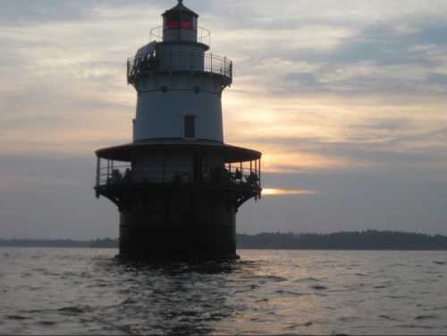 A lighthouse stands in the water at sunset, with a cloudy sky and distant trees visible on the horizon.