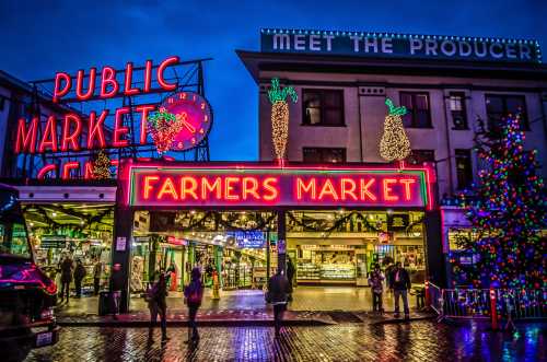 Brightly lit farmers market entrance with festive decorations, surrounded by shops and a colorful Christmas tree.