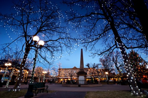 A festive park scene at dusk, adorned with twinkling lights and a central obelisk, surrounded by trees and a Christmas tree.