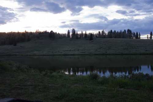 A serene landscape featuring a calm lake reflecting the cloudy sky and a grassy hillside with trees in the background.