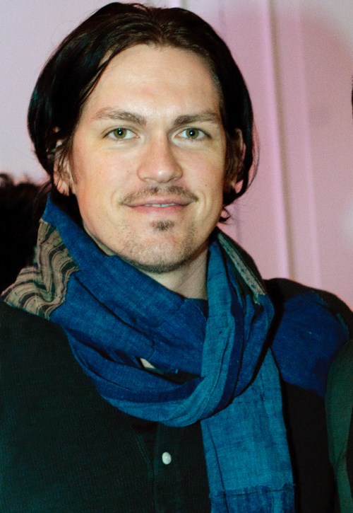 A man with long dark hair and a blue scarf smiles at the camera, wearing a black shirt.