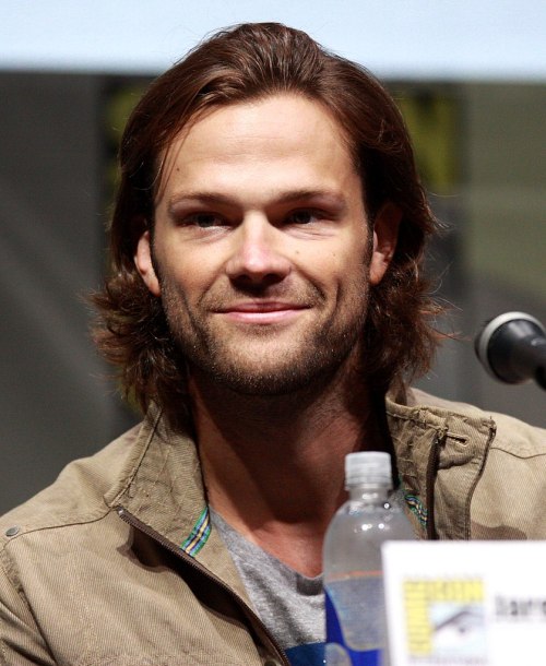 A man with long hair smiles at a panel, wearing a casual jacket and sitting in front of a water bottle.