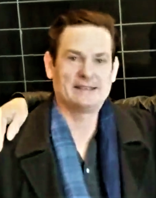 A man with short hair wearing a black coat and blue scarf, smiling in front of a dark tiled background.