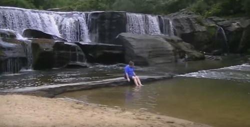 A person sits on a rock by a serene waterfall, with water flowing gently around them and a sandy shore nearby.