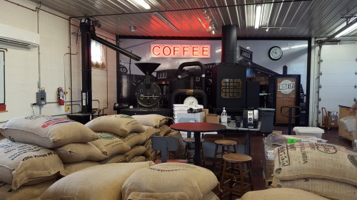 A coffee roasting facility with burlap sacks, a roasting machine, and a neon "COFFEE" sign in the background.