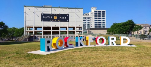 Colorful "ROCKFORD" sign in front of Davis Park, with a clear blue sky and modern buildings in the background.