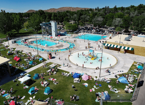 Aerial view of a busy public pool area with multiple pools, sunbathers, and colorful umbrellas on a sunny day.