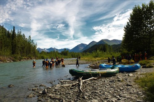 Group of people preparing for rafting on a river surrounded by mountains and trees under a blue sky.