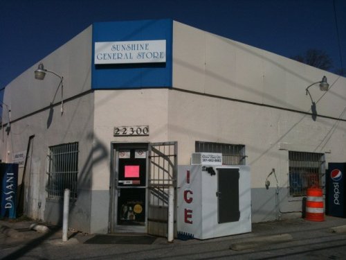 Exterior of Sunshine General Store, featuring a blue sign, ice vending machine, and a white building with barred windows.