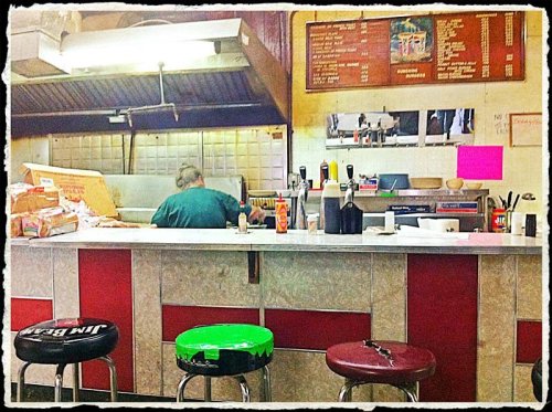 A retro diner counter with three stools, a cook in the back, and a menu board on the wall.