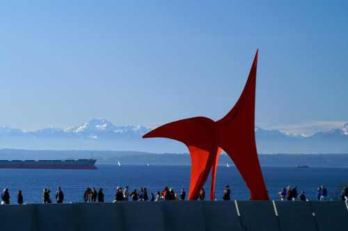 A large red sculpture stands by the water, with people walking nearby and mountains in the background under a clear sky.