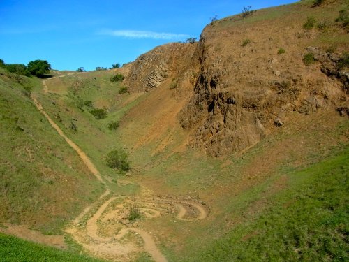 A winding dirt path leads through a dry, hilly landscape with sparse vegetation under a clear blue sky.