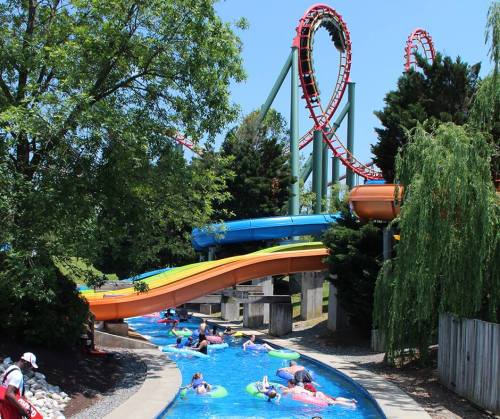 A water park scene with a winding slide and a roller coaster in the background, featuring people floating in a lazy river.