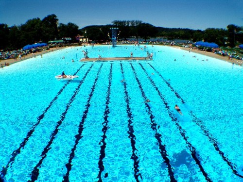 A large outdoor swimming pool with clear blue water, lanes marked, and people enjoying the sun and swimming.