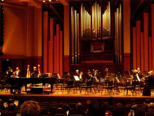 A concert hall stage set up with a grand piano, orchestra seating, and a large pipe organ in the background.