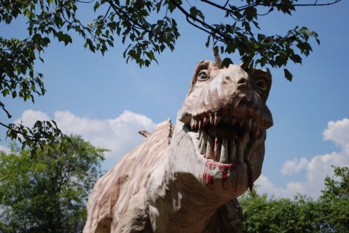 A large, realistic dinosaur sculpture with sharp teeth, set against a blue sky and green trees.