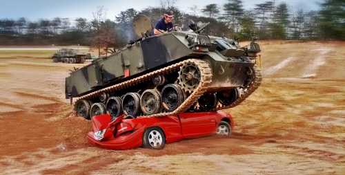 A tank drives over a crushed red car in a dirt field, showcasing the tank's power and size.