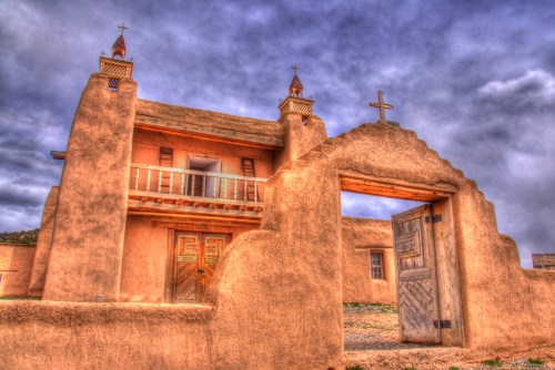 Historic adobe building with a wooden door and crosses, set against a dramatic cloudy sky.