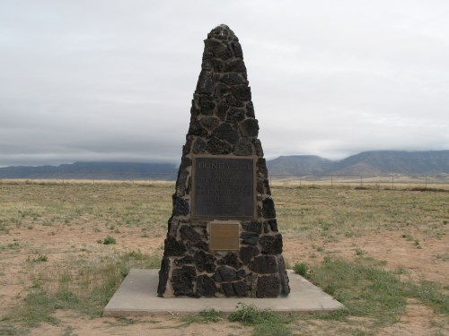 A tall stone monument with plaques stands in a grassy area, surrounded by a flat landscape and distant mountains.
