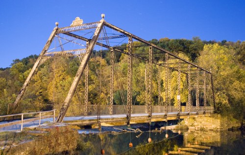 A historic metal bridge spans a calm river, surrounded by trees with autumn foliage under a clear night sky.