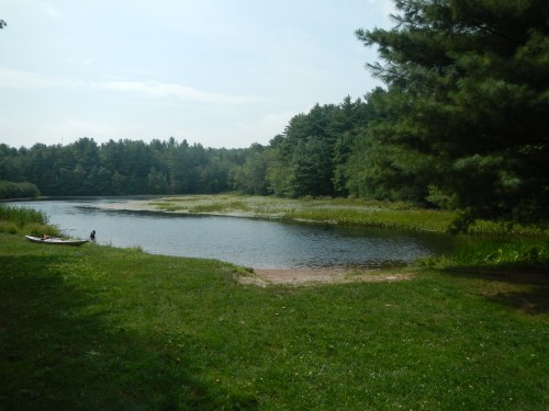 A serene lake surrounded by lush greenery and trees, with a small sandy area and a person near the water's edge.
