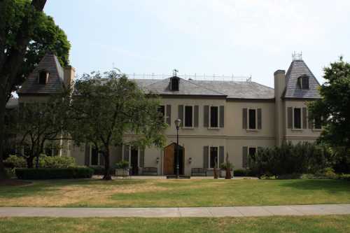 A large, elegant mansion with a slate roof, surrounded by trees and a well-maintained lawn on a sunny day.