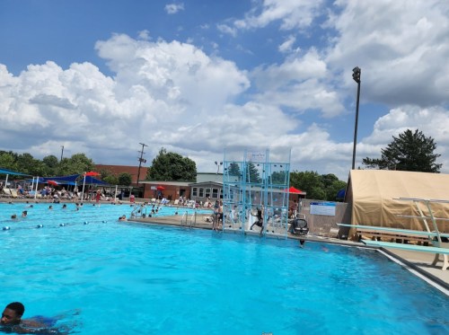 A sunny outdoor pool scene with people swimming, diving boards, and colorful umbrellas under a partly cloudy sky.