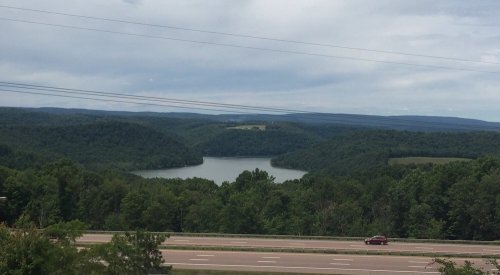 A scenic view of a lake surrounded by green hills under a cloudy sky, with a highway in the foreground.
