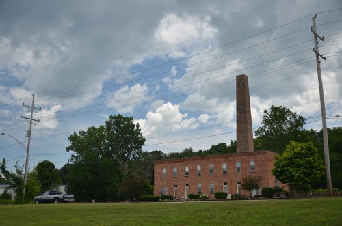 A brick building with a tall chimney, surrounded by trees and power lines, under a cloudy sky.