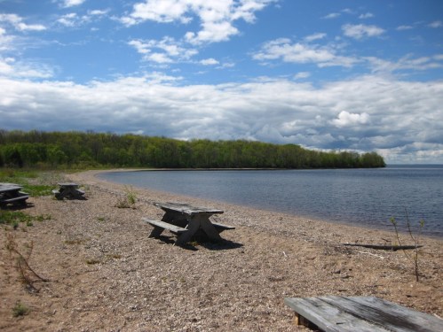A serene beach scene with picnic tables, sandy shore, and lush green trees under a partly cloudy sky.