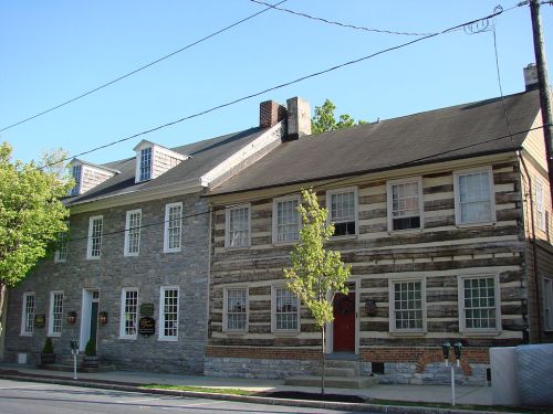 Two historic buildings side by side: one stone and one log cabin style, with trees and a clear blue sky in the background.