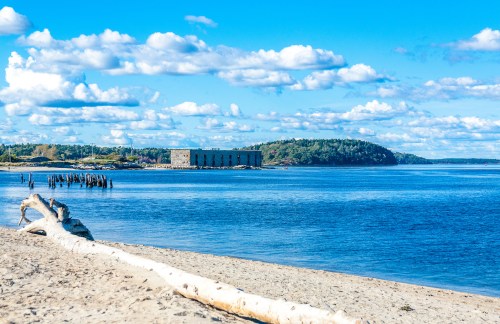 A sandy beach with a log, calm blue water, and a distant historic building under a bright, cloudy sky.