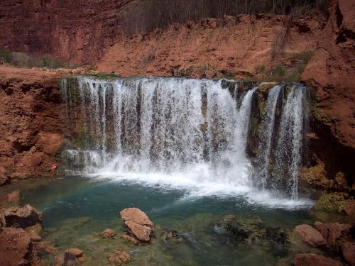 A cascading waterfall flows into a clear pool, surrounded by red rock formations and lush greenery.