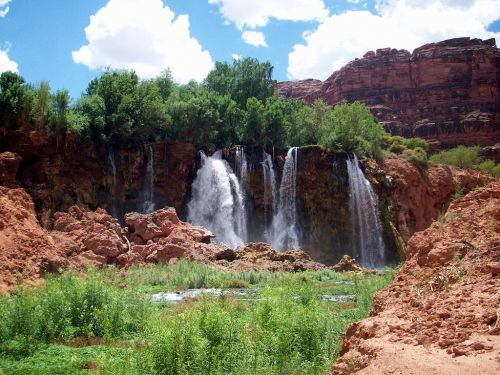 A scenic waterfall cascading over red rock formations, surrounded by lush greenery and blue skies with fluffy clouds.