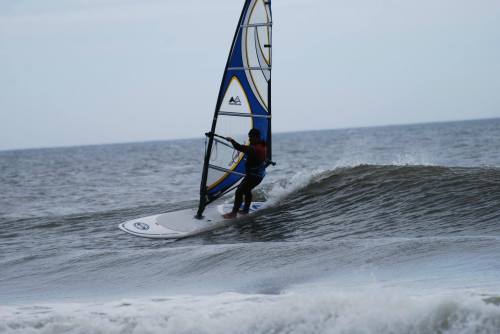 A windsurfer glides over a wave on a cloudy day, with a blue and yellow sail catching the wind.