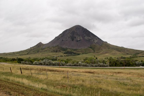 A large, conical mountain rises above a grassy landscape under a cloudy sky, with trees lining the foreground.
