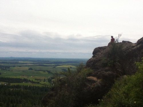 A person sits on a rocky outcrop, overlooking a vast green landscape under a cloudy sky.