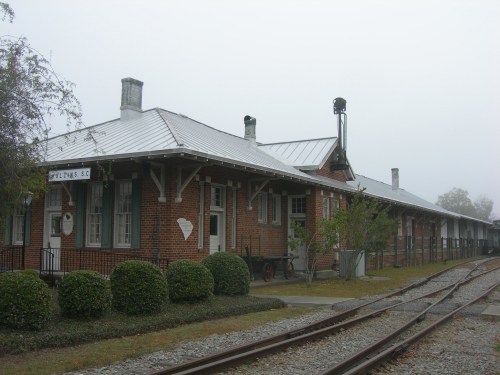 Historic train station with brick architecture, metal roof, and foggy surroundings, featuring nearby railway tracks.