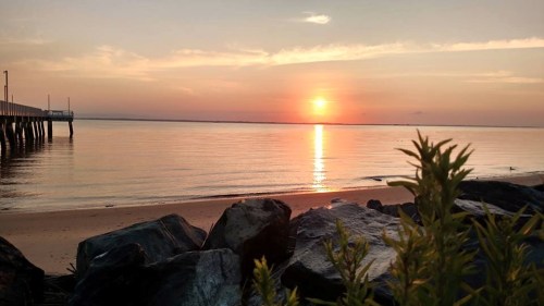Sunset over calm waters, with a pier in the distance and rocks in the foreground, creating a serene coastal scene.