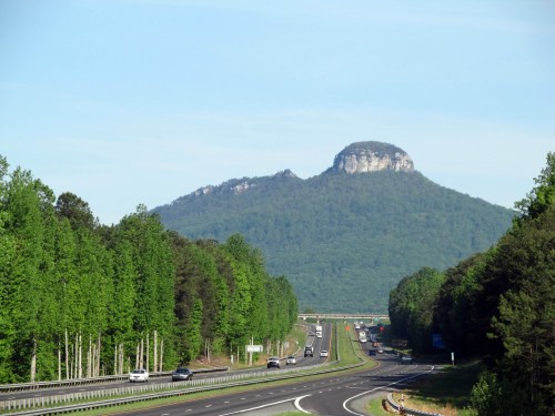 A highway stretches towards a mountain peak, surrounded by lush green trees under a clear blue sky.