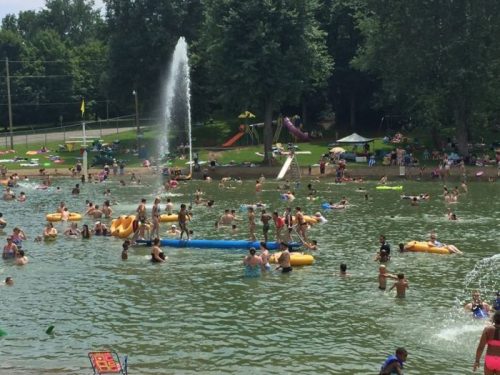 A busy swimming area with people on floats, a fountain, and playground equipment in the background on a sunny day.
